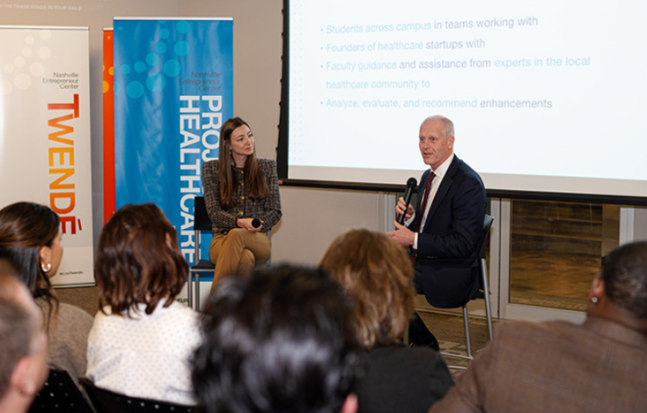 Vanderbilt Business Dean Steenburgh speaks on the value of experimental learning to the audience. Steenburgh is sitting at the front of the room in front of a slide deck presentation and is holding a microphone.