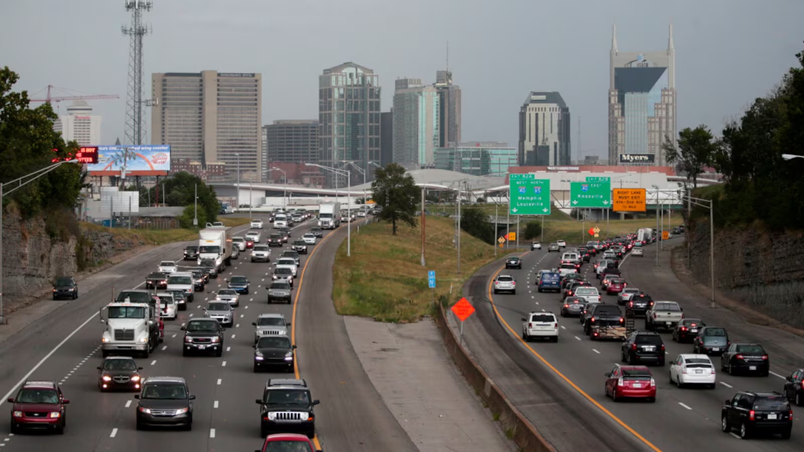 View of a busy highway in Nashville, Tennessee, with multiple lanes of traffic in both directions. The downtown skyline is visible in the background, featuring prominent buildings such as the AT&T 'Batman' Building. Road signs point to destinations like Memphis, Louisville, and Knoxville