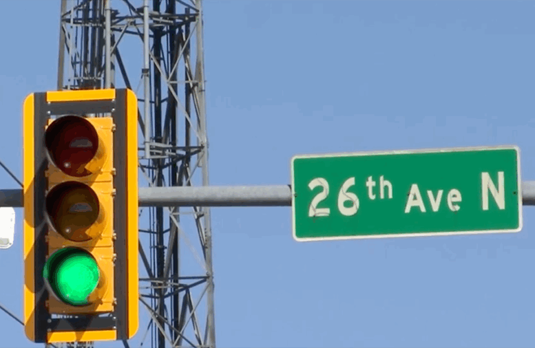 Traffic light showing green signal next to a street sign for 26th Avenue North under clear blue sky.
