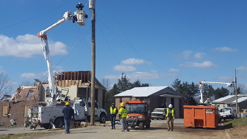 Utility crews repair power lines near tornado-damaged homes while workers assess the scene and equipment is staged for cleanup.