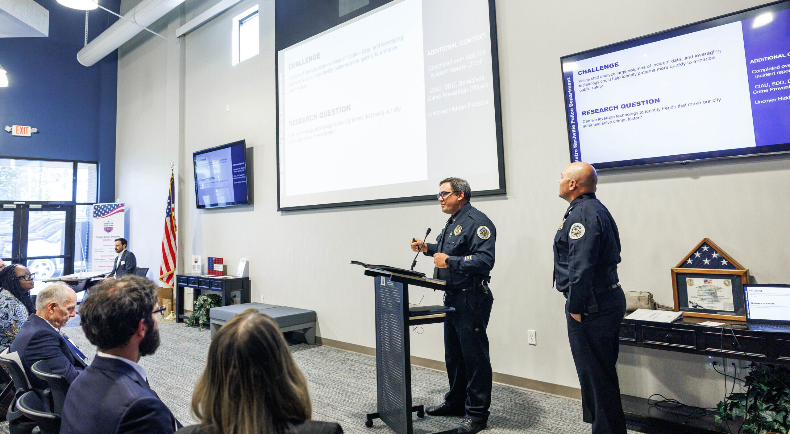 alt="Two police officers present tech challenges to an audience in a conference room with screens displaying slides and an American flag."