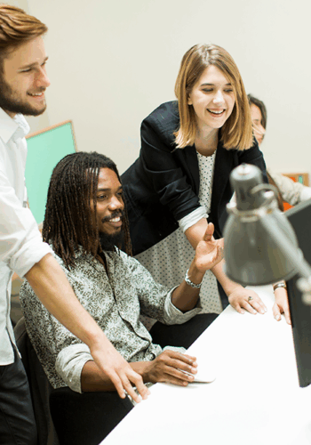 Three colleagues collaborate around a desk with computers, under modern lamps, in a bright office setting.