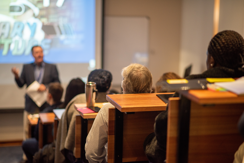 Students sit in tiered seating listening to a presenter speaking in front of a projected screen.