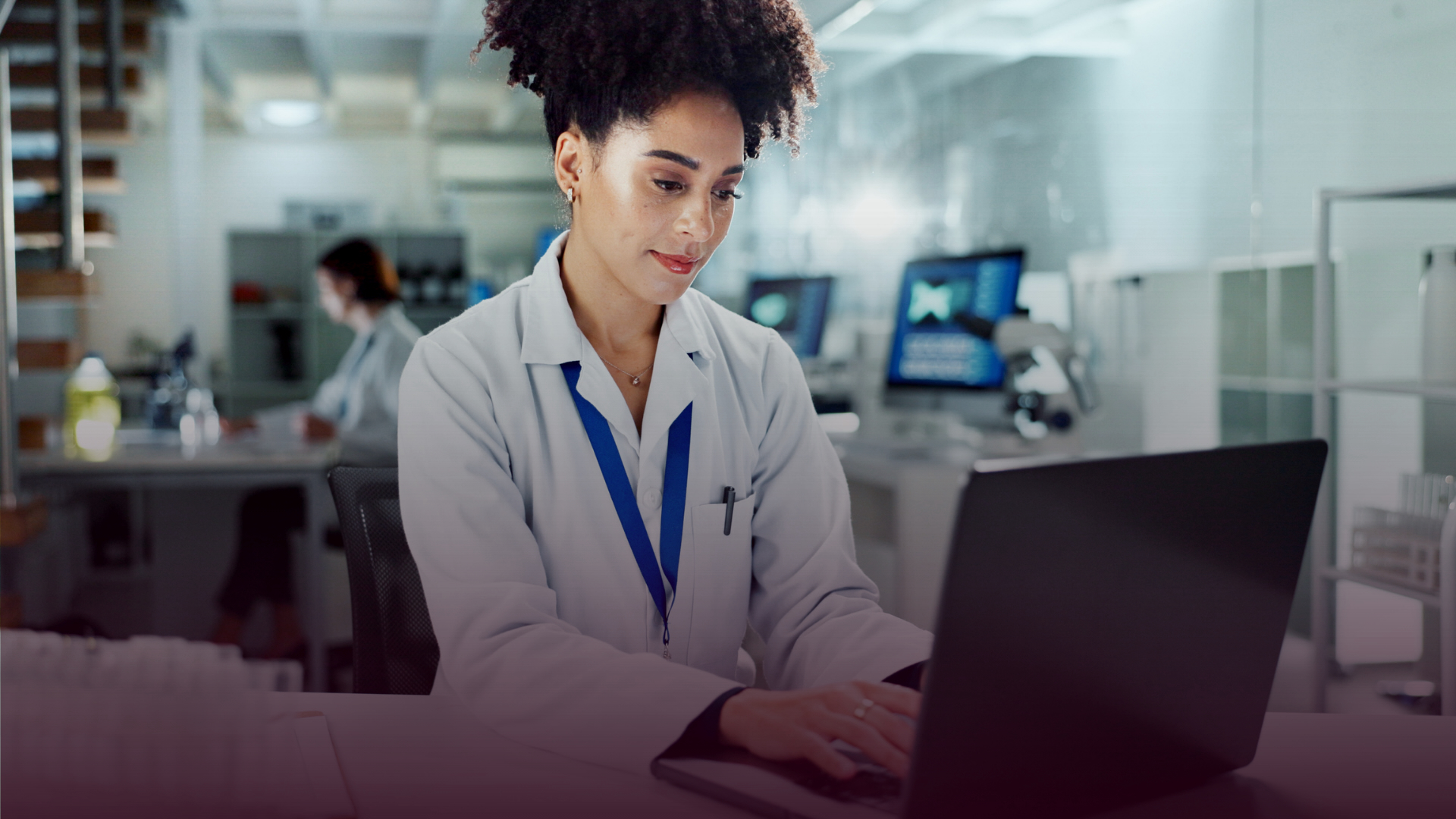 Scientist in a lab coat working on a laptop in a laboratory, with equipment and monitors in the background.