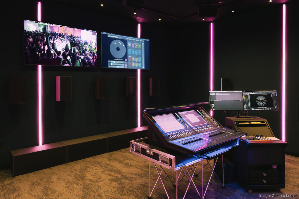 Audio production studio with mixing console, monitors, wall-mounted speakers, and pink accent lighting.