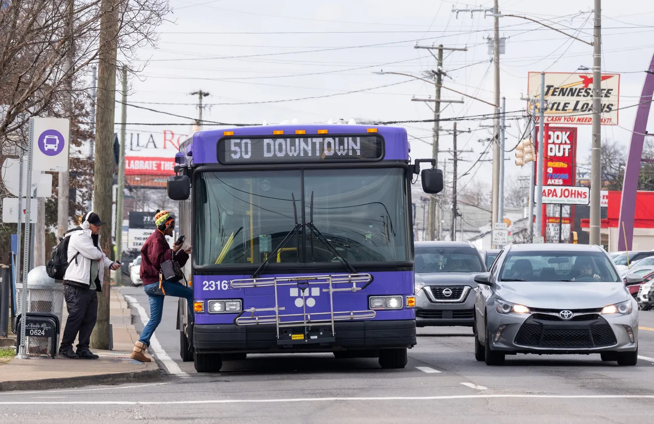 City bus labeled “50 Downtown” stops to pick up passengers at a roadside bus stop with cars passing nearby.