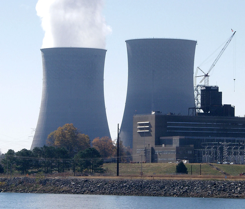 Nuclear power plant with two large cooling towers releasing steam beside industrial buildings near a body of water.